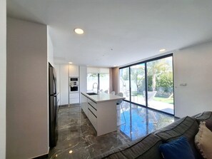 A modern kitchen with a central island, a dining area with a glass table, and a large sliding glass door leading to an outdoor space. - Panorama Luxury Summer Villa (Paralimni)