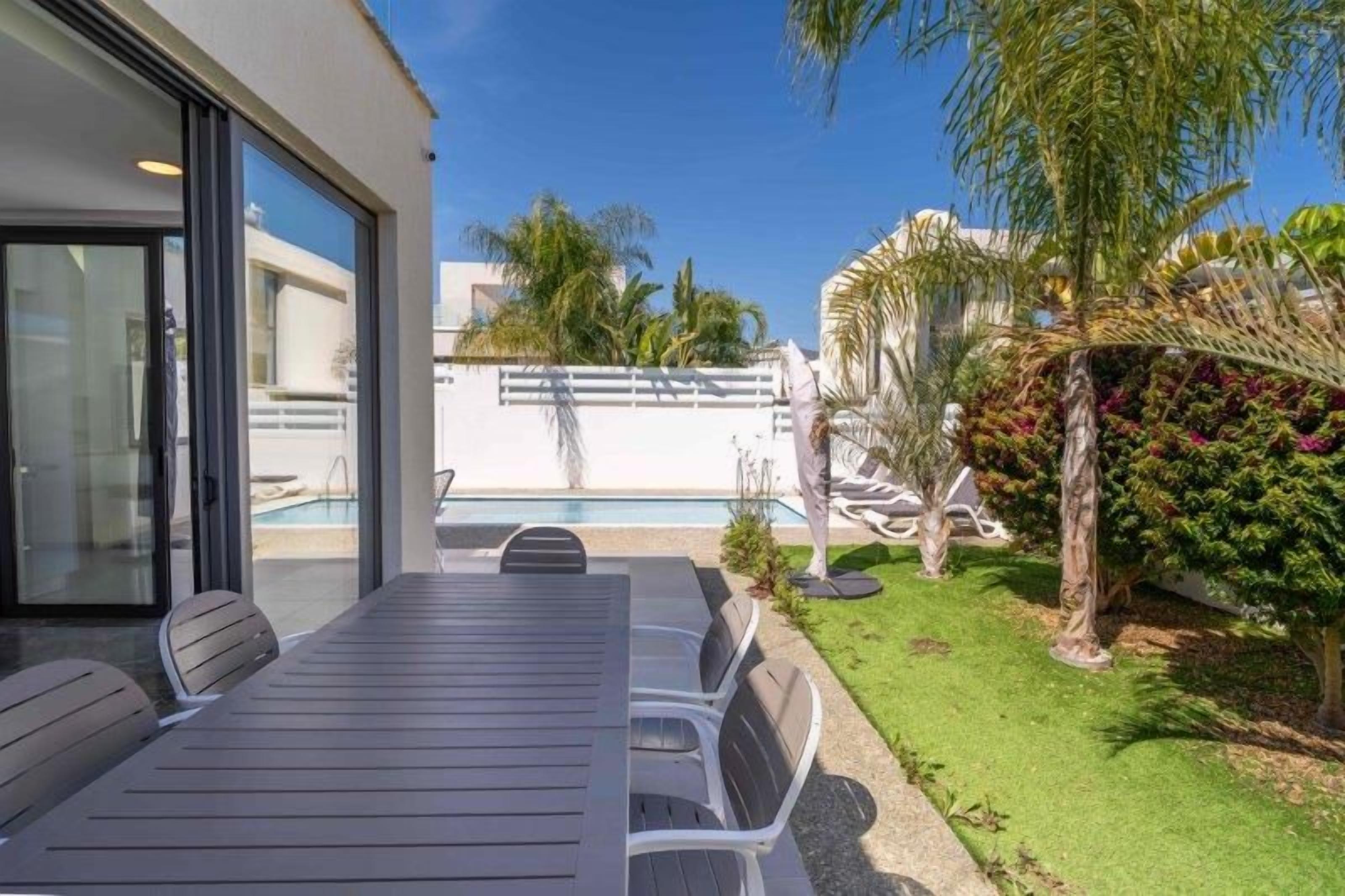 A modern outdoor patio with a dining table and chairs, a pool, and a well-maintained lawn with palm trees.