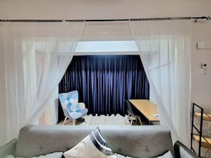 Ein Zimmer mit einem Bett, einem Sessel, einem Tisch und Blick durch ein Fenster mit Vorhängen.