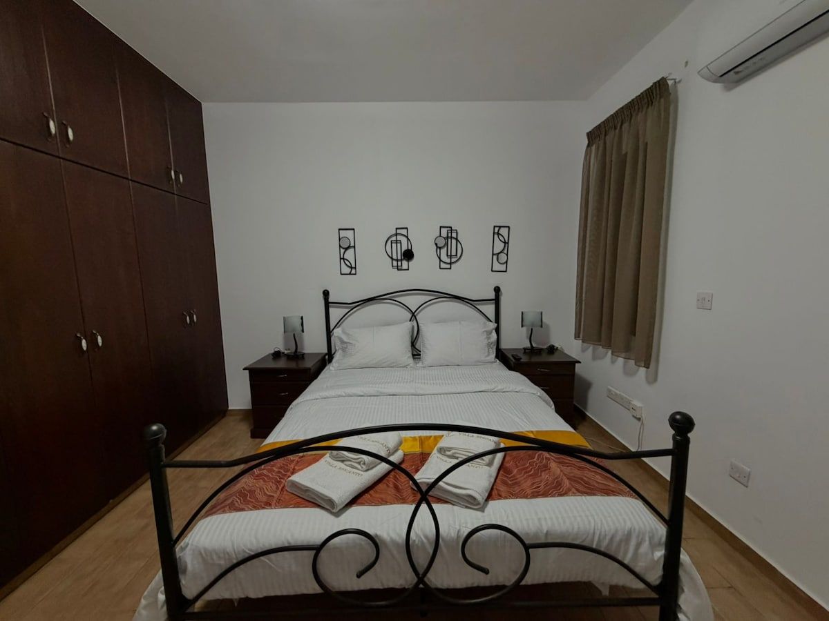 A bedroom with a bed, wooden wardrobe, bedside tables, and a wall-mounted air conditioner.