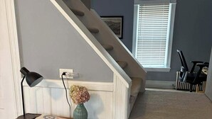 Eine Treppe mit Holzvertäfelung, ein kleiner Holztisch mit einer Vase und einer Lampe sowie ein Fenster mit Rollläden.