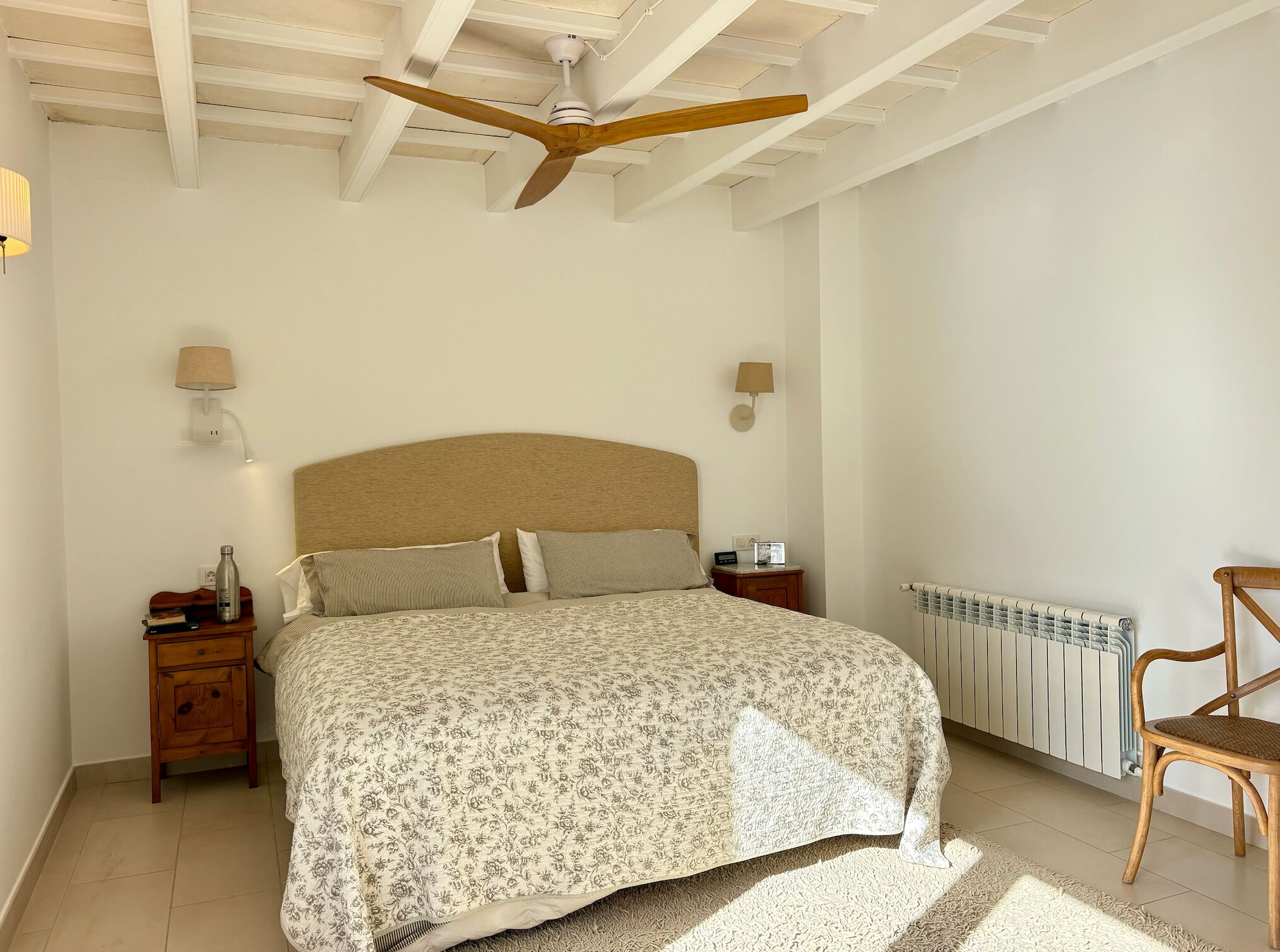 A bedroom with a bed, wooden headboard, bedside tables, lamps, a radiator, and a ceiling fan.