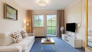 A hotel room with a sofa, a coffee table, a TV, a fan, and a window with a view of greenery.