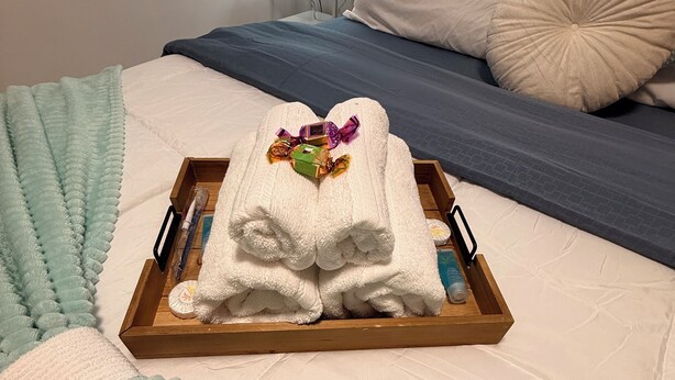 Una cama bien hecha con una colcha a rayas azul y blanco, una almohada blanca y una bandeja de madera con toallas dobladas y un pequeño adorno.