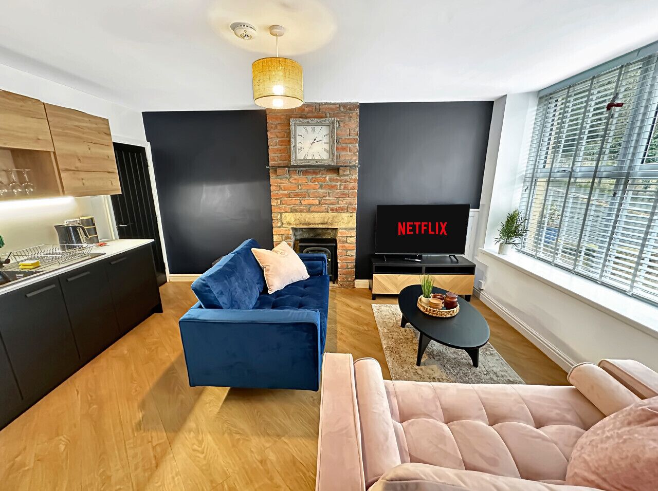 Un salon moderne avec un canapé bleu, un fauteuil rose, une cheminée, un téléviseur et un coin cuisine.