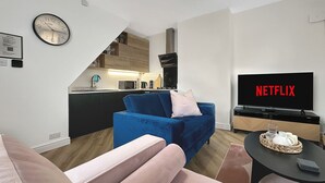 En moderne stue med en blå sofa, en pink sofa, et fjernsyn der viser 'NETFLIX' og et køkken i baggrunden.