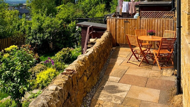 En stenbelagt terrasse med et bord og stole, et træhegn og udsigt til huse og grønne områder.