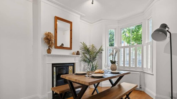 A dining area with a wooden table and benches, a fireplace, and a large window with shutters. - Relaxed Family Retreat in Kingston (Kingston Upon Thames)