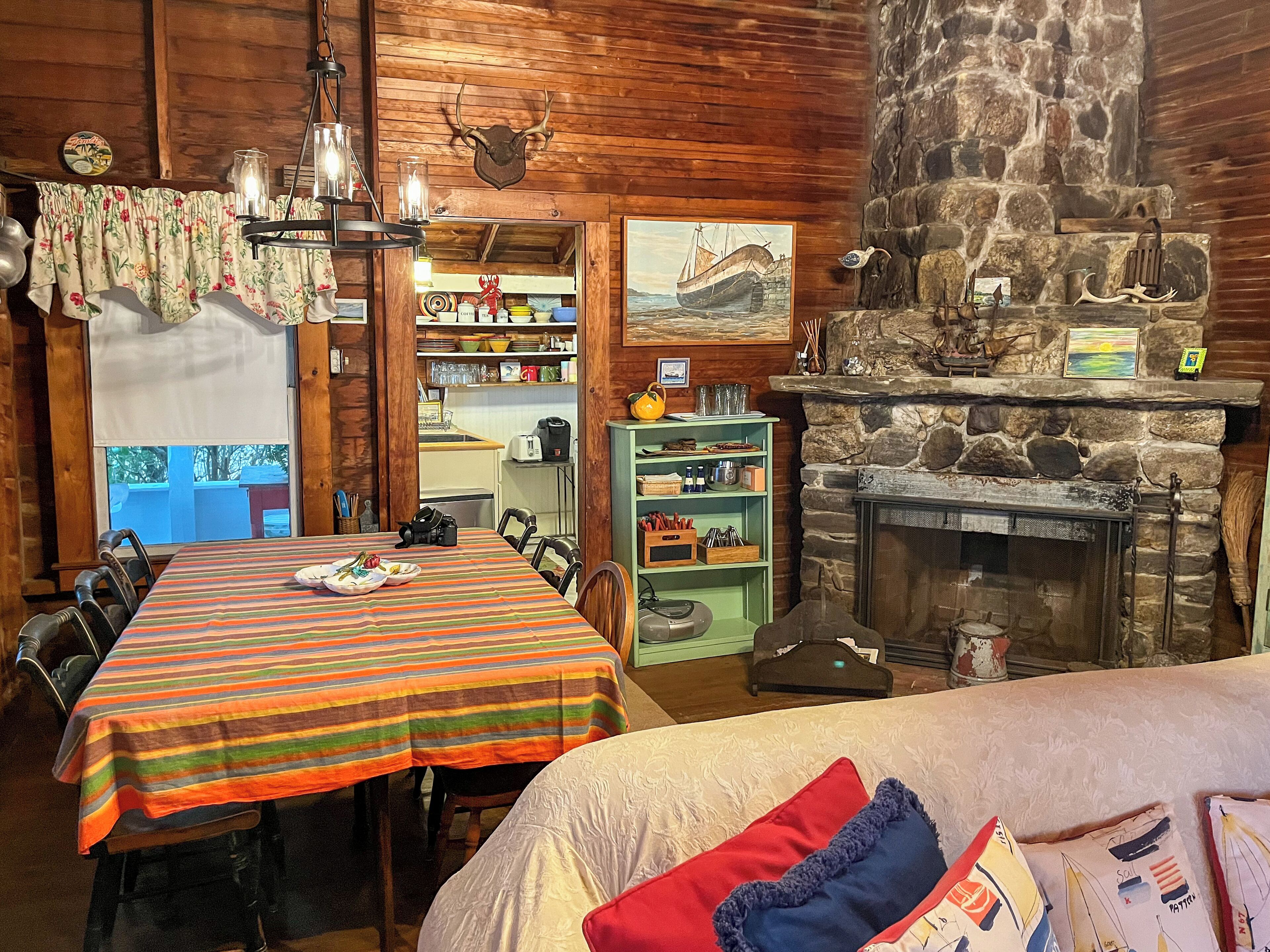 A cozy cabin interior with a dining table, chairs, a fireplace, and a kitchen area visible through an open door.