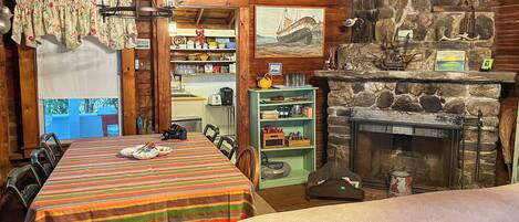A cozy cabin interior with a dining table, chairs, a fireplace, and a kitchen area visible through an open door.