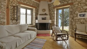 Une pièce aux murs de pierre, dotée d’une cheminée, de poutres en bois au plafond, d’un canapé et de deux fauteuils à bascule.