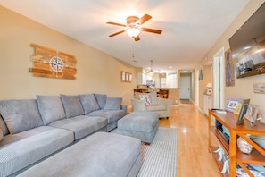 A living room with a sectional sofa, a ceiling fan, a kitchen in the background, and a bookshelf with various items. - Fantastic True Blue 2 bedroom golf front condo (Pawleys Island)
