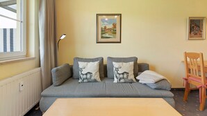 Ein Zimmer mit grauem Sofa, zwei Kissen mit Hirschmotiv, einem gerahmten Landschaftsbild, einem Holzstuhl und einem Fenster mit Vorhängen.