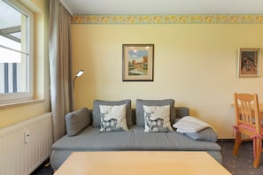 Ein Zimmer mit grauem Sofa, zwei Kissen mit Hirschmotiv, einem gerahmten Landschaftsbild, einem Holzstuhl und einem Fenster mit Vorhängen.