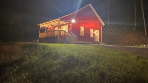 Una cabaña roja de madera con un porche y un espacio cubierto, rodeada de árboles.