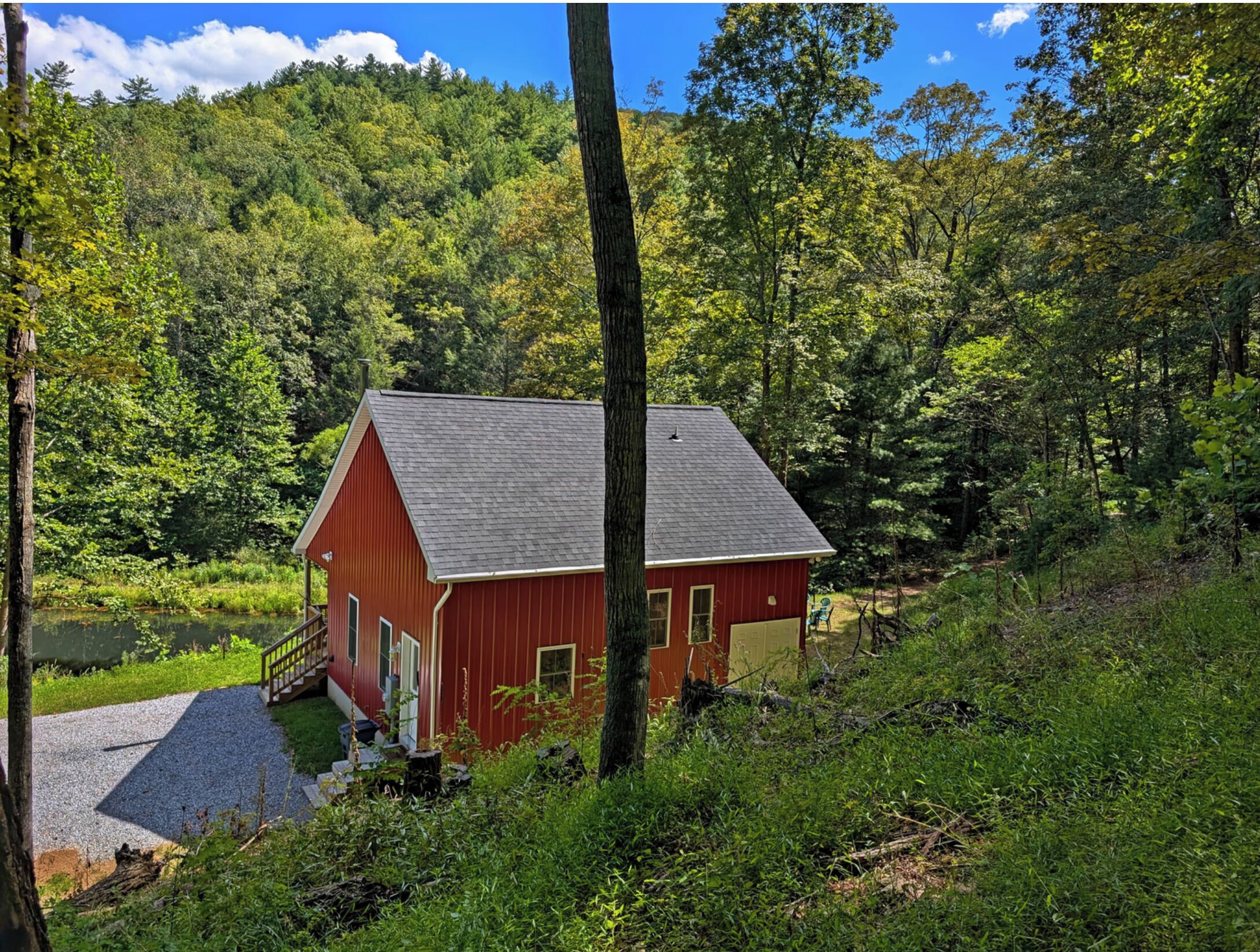 A red cabin with a gravel driveway, surrounded by dense forest and a hill in the background.