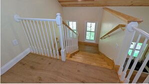 Una escalera de madera con barandillas blancas, un techo con vigas de madera y vistas al verdor exterior.