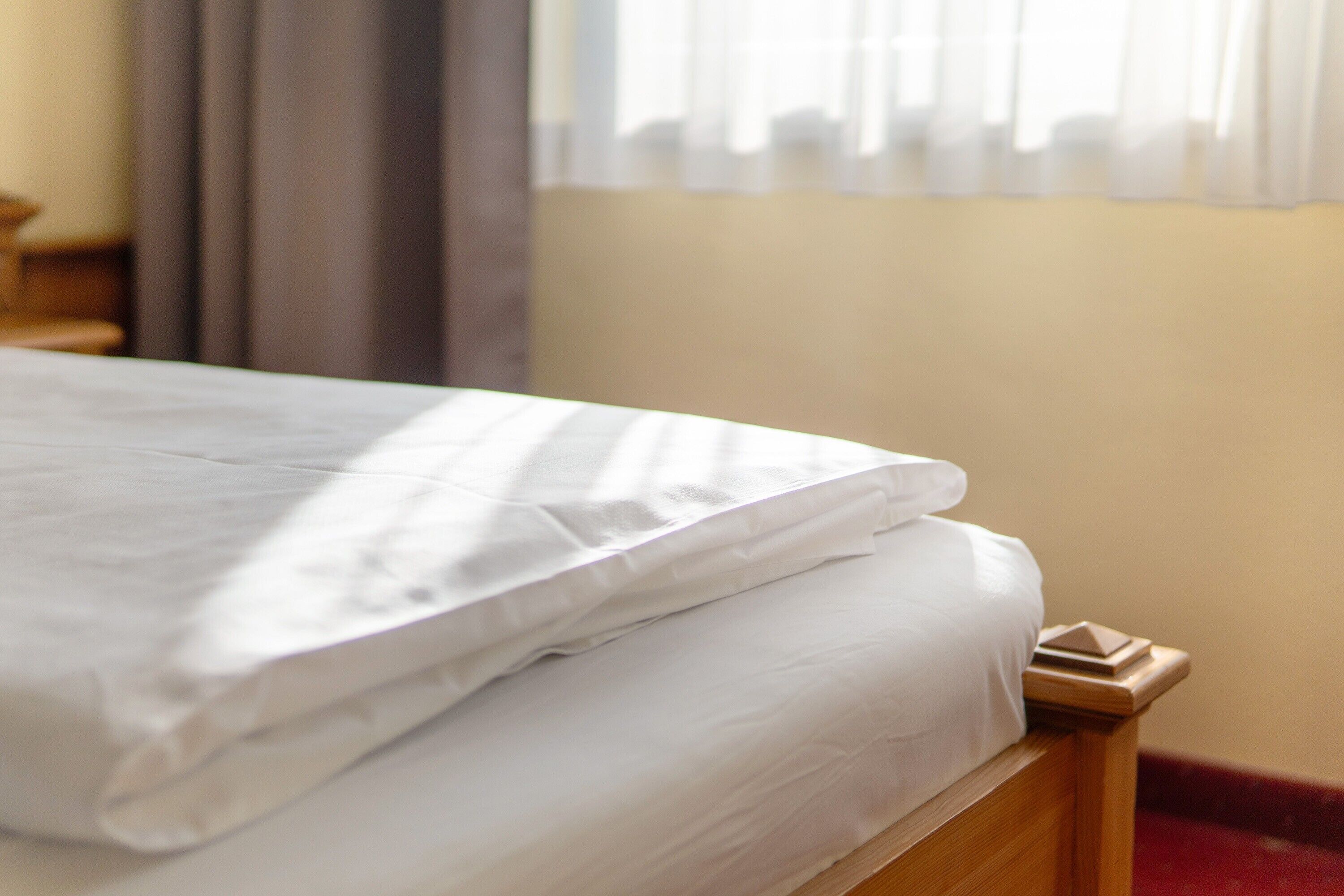 Una cama bien tendida con sábanas blancas, cabecero de madera y una ventana con cortinas traslúcidas.