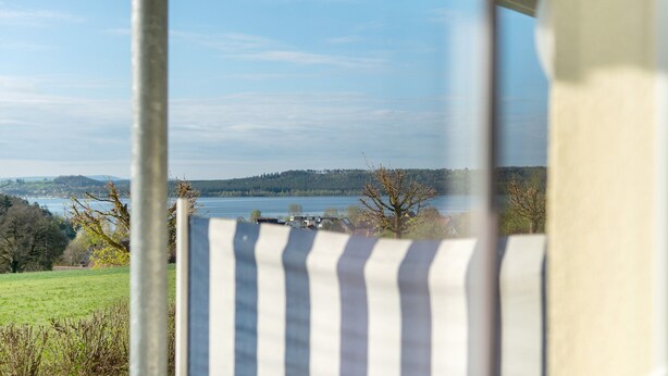 Une vue à travers une fenêtre, avec un poteau métallique et un objet rayé au premier plan, un lac et une ligne d’arbres à l’horizon.