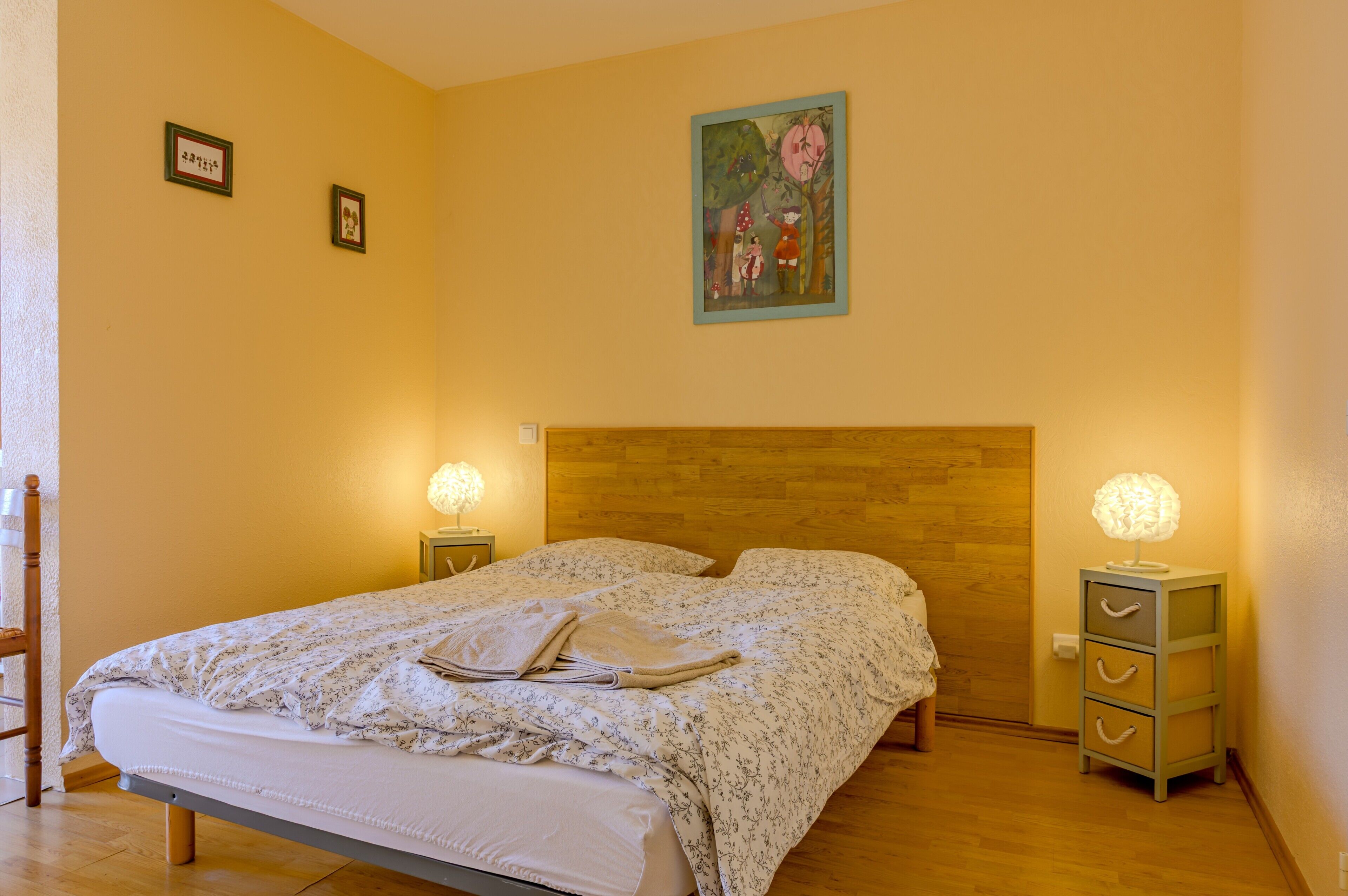 Une chambre à coucher avec une tête de lit en bois, un lit recouvert d’une couvre-lit à motifs floraux, deux tables de chevet avec des lampes et une petite commode à tiroirs.