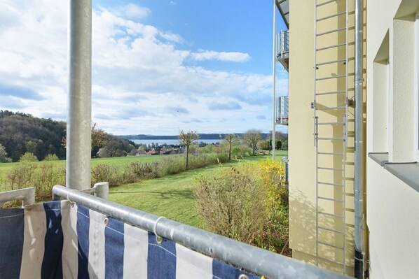 A balcony with a metal railing, a view of a green landscape, and a building with a yellow exterior.