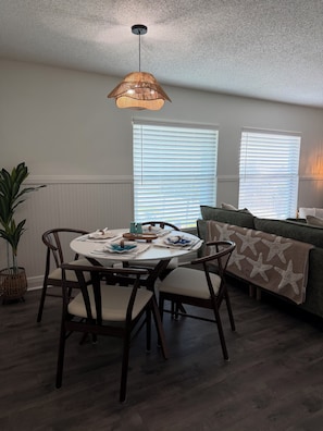 Un coin repas avec une table ronde, des chaises et un luminaire suspendu. Il y a deux fenêtres équipées de stores, une plante en pot et un canapé agrémenté d’un plaid à motifs d’étoiles de mer.