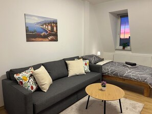 Un dormitorio moderno con un sofá gris, una cama con colcha estampada, una mesita de madera y una ventana con vistas al atardecer.