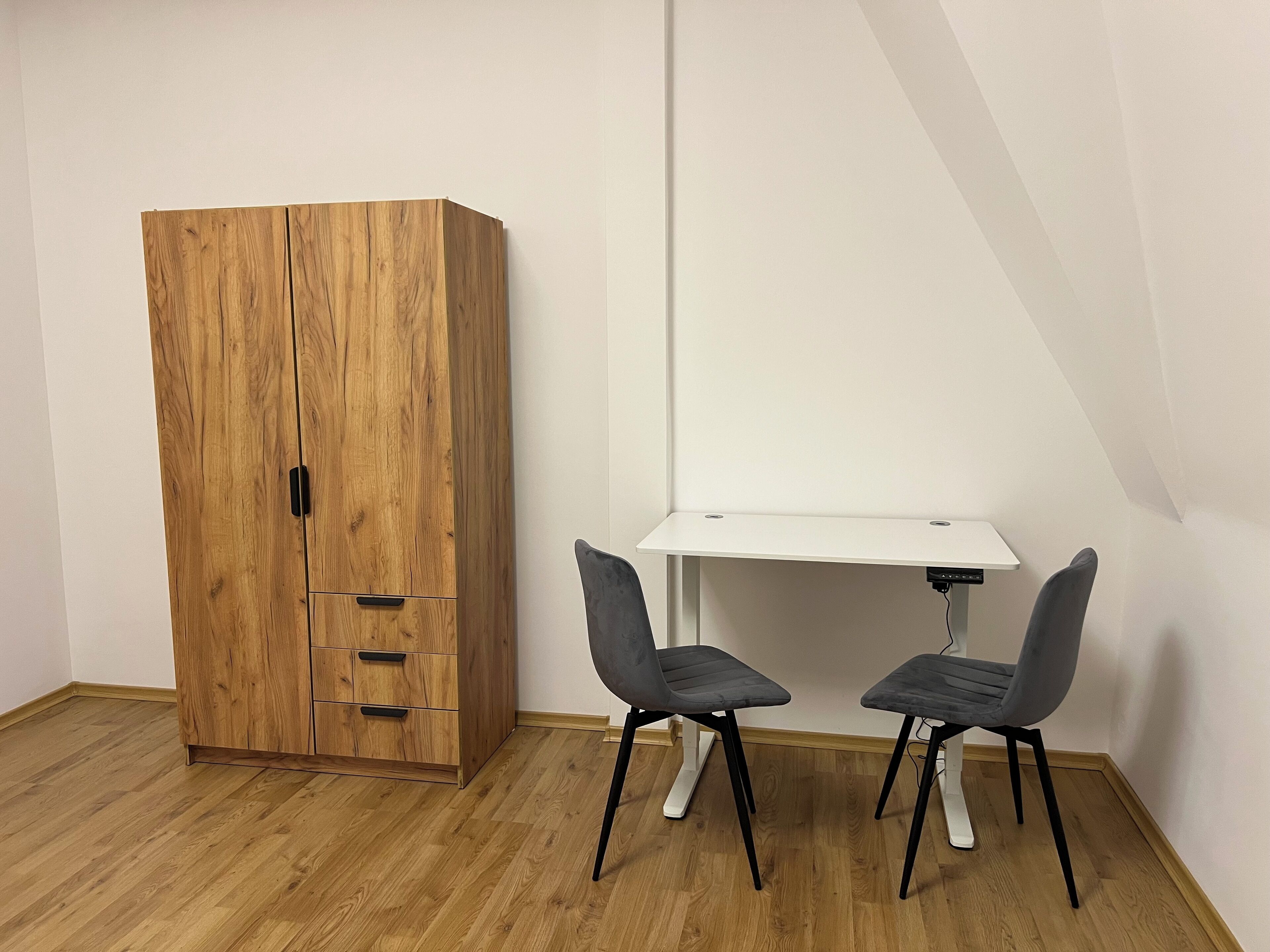 A room with a wooden wardrobe, a white desk, and two grey chairs.