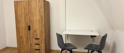 A room with a wooden wardrobe, a white desk, and two grey chairs.
