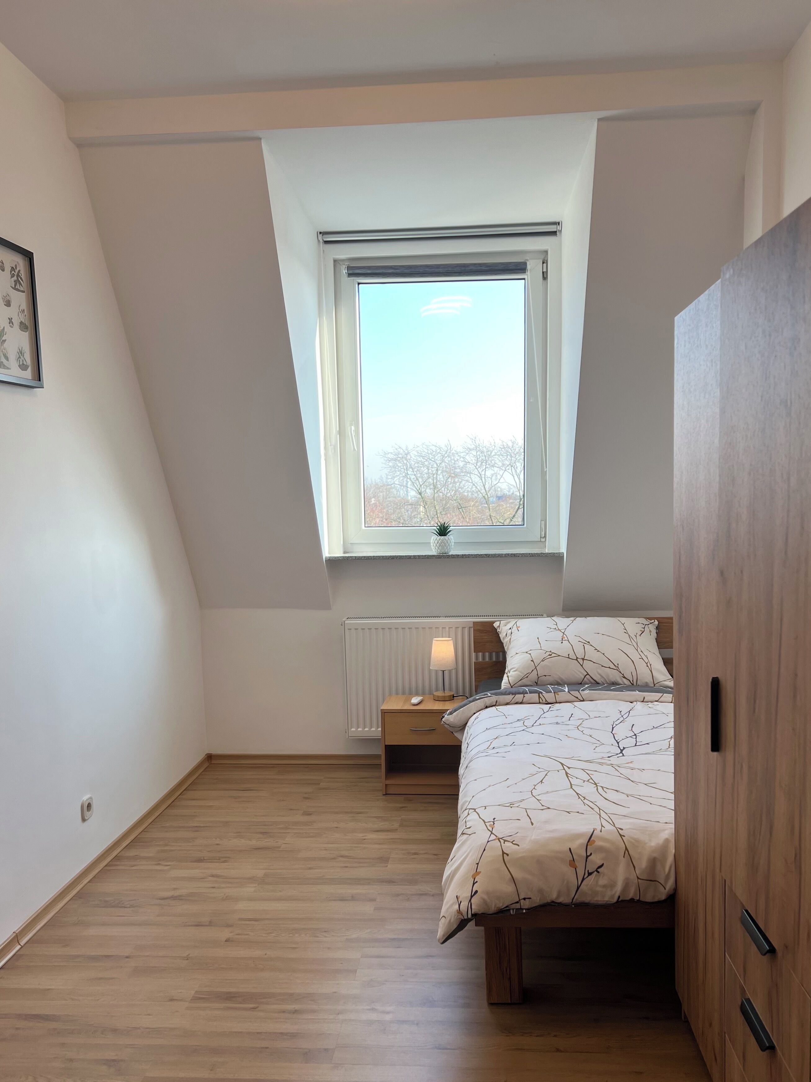 Une chambre avec un plafond en pente, une fenêtre donnant sur les arbres, une table de chevet avec une lampe et un lit recouvert d’une couette à motifs de branches.