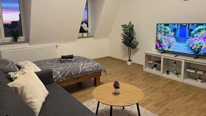 En moderne stue med en seng, en sofa, et rundt bord, en potteplante, et fjernsyn og et vindue med udsigt til solnedgang.