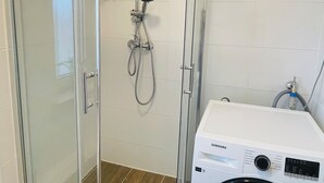 Et brusebad med glasvægge, en håndbruser og en vandhane på væggen. En Samsung vaskemaskine er synlig til højre.