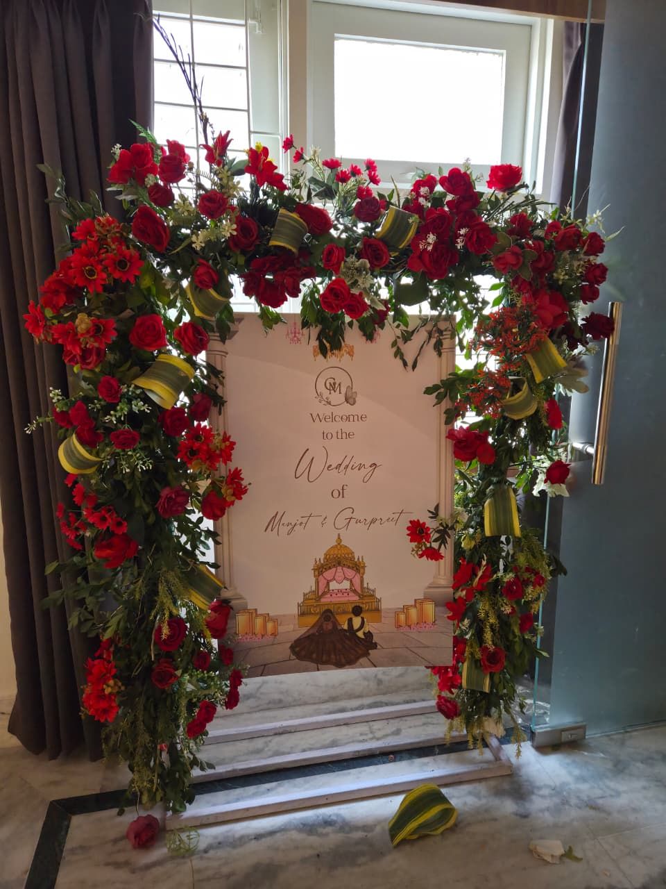 A wedding arch with red flowers and a welcome sign for the wedding of Minjit & Gurpreet.