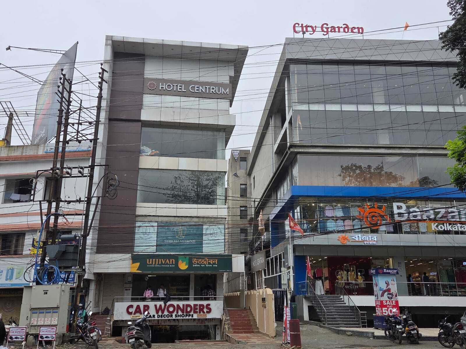 A hotel named Hotel Centrum with a modern glass facade, adjacent to a shopping area with visible stores like Baazar and a car decor shop named Cap Wonders.