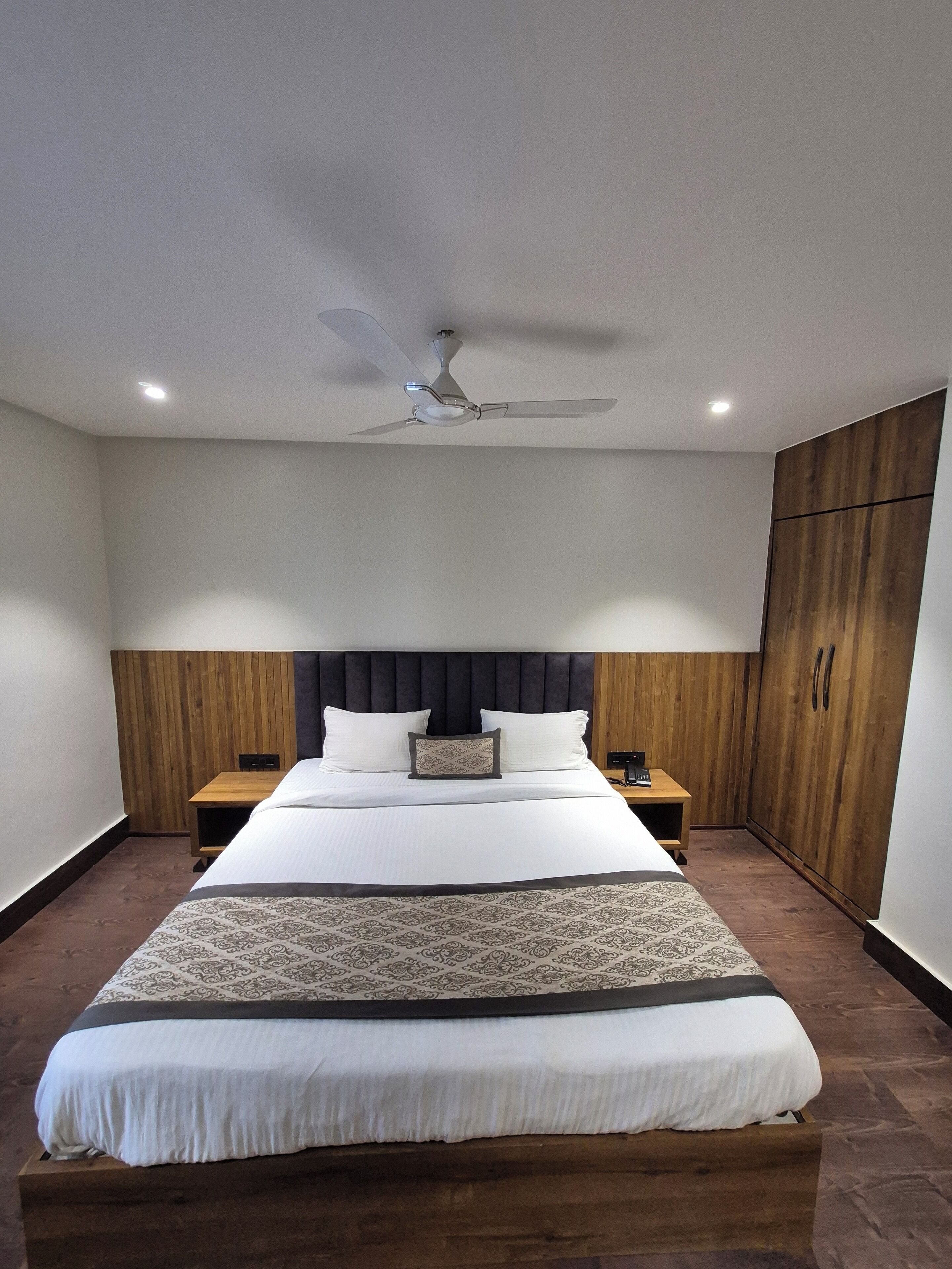 A modern bedroom with a large bed, wooden headboard, two bedside tables, a ceiling fan, and a wardrobe.
