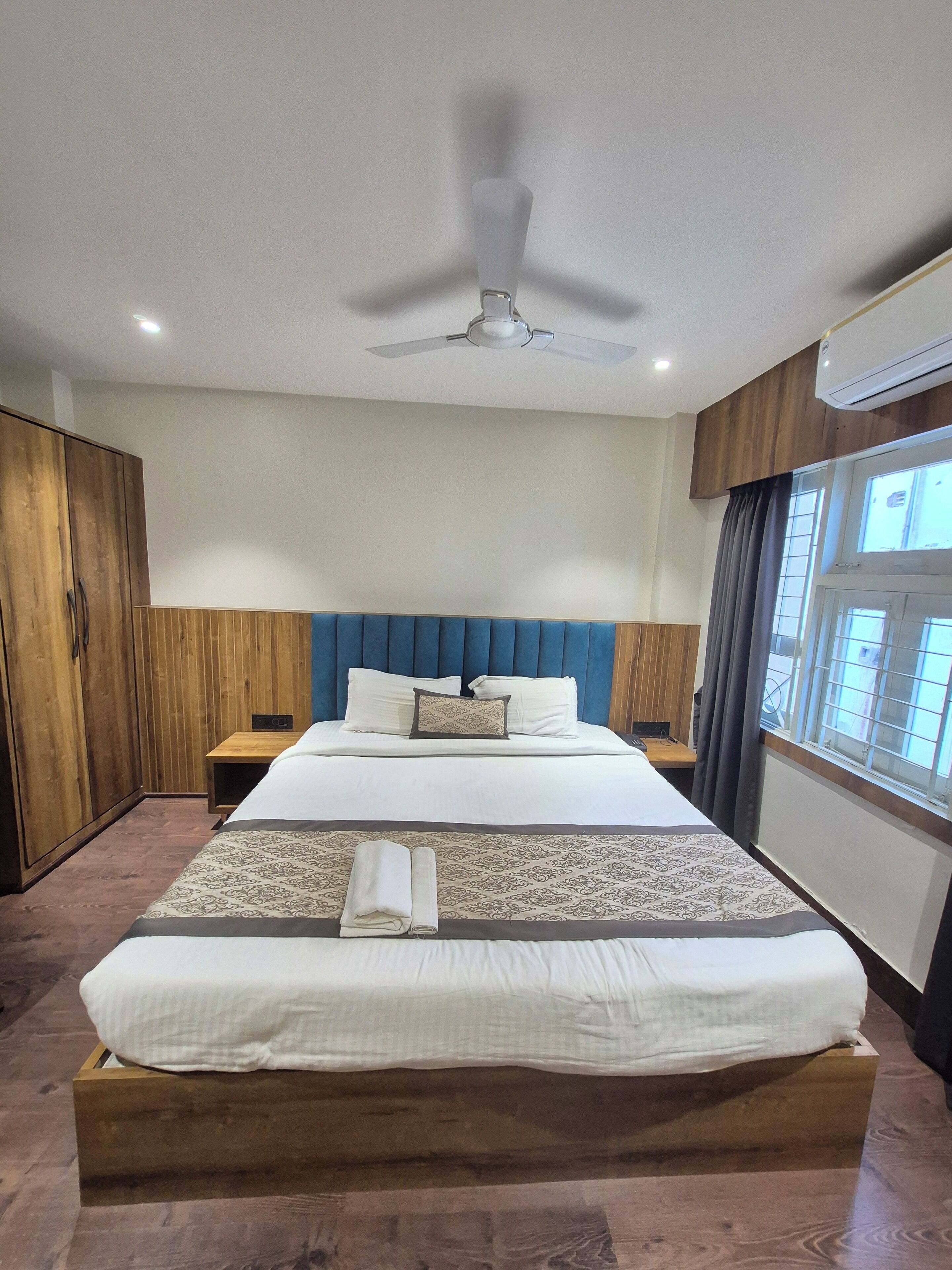 Habitación de hotel con una cama grande, cabecero de madera, ventilador de techo y ventana con cortinas.