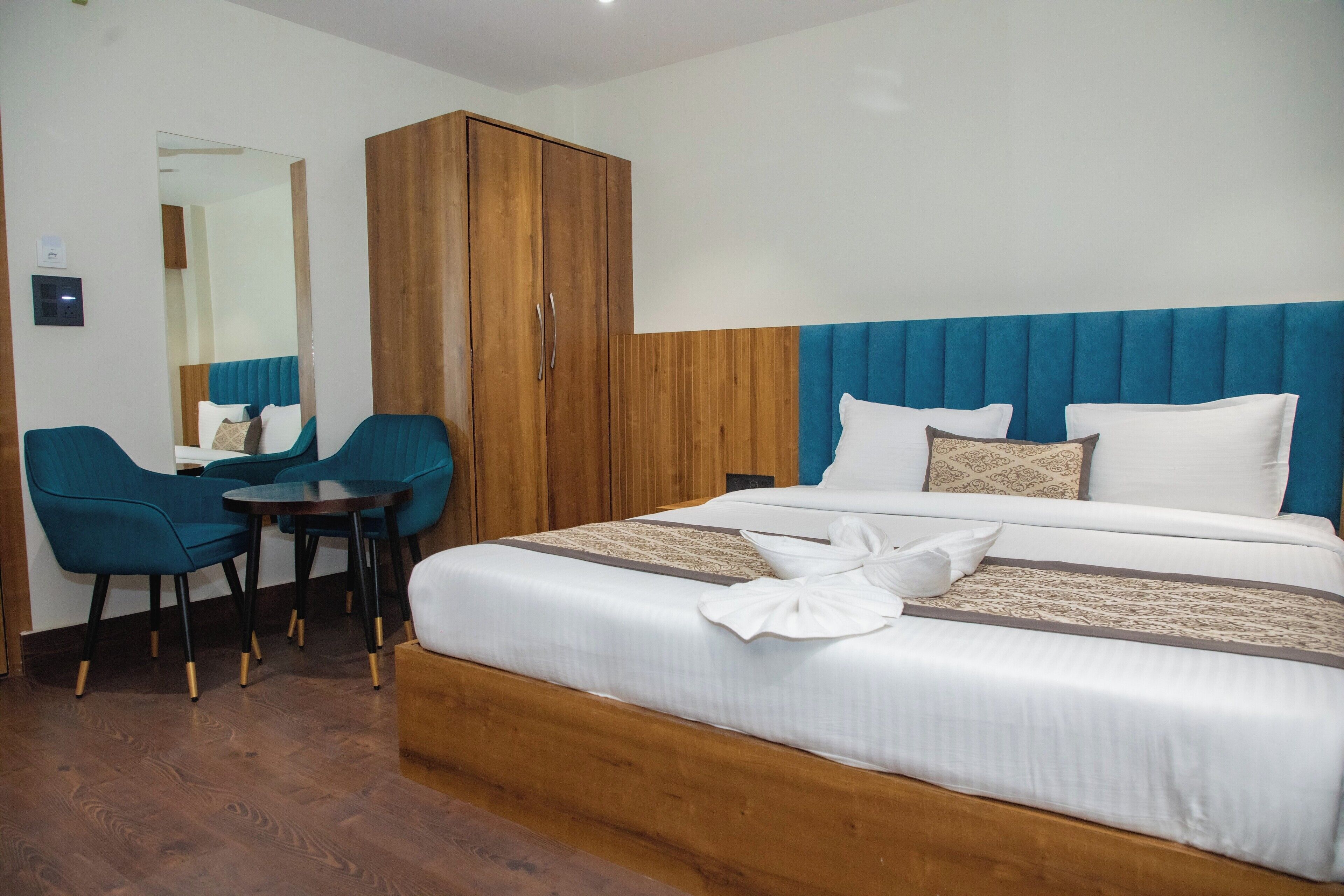 A hotel room with a large bed, a wooden wardrobe, a small table, two blue chairs, and a mirror on the wall.