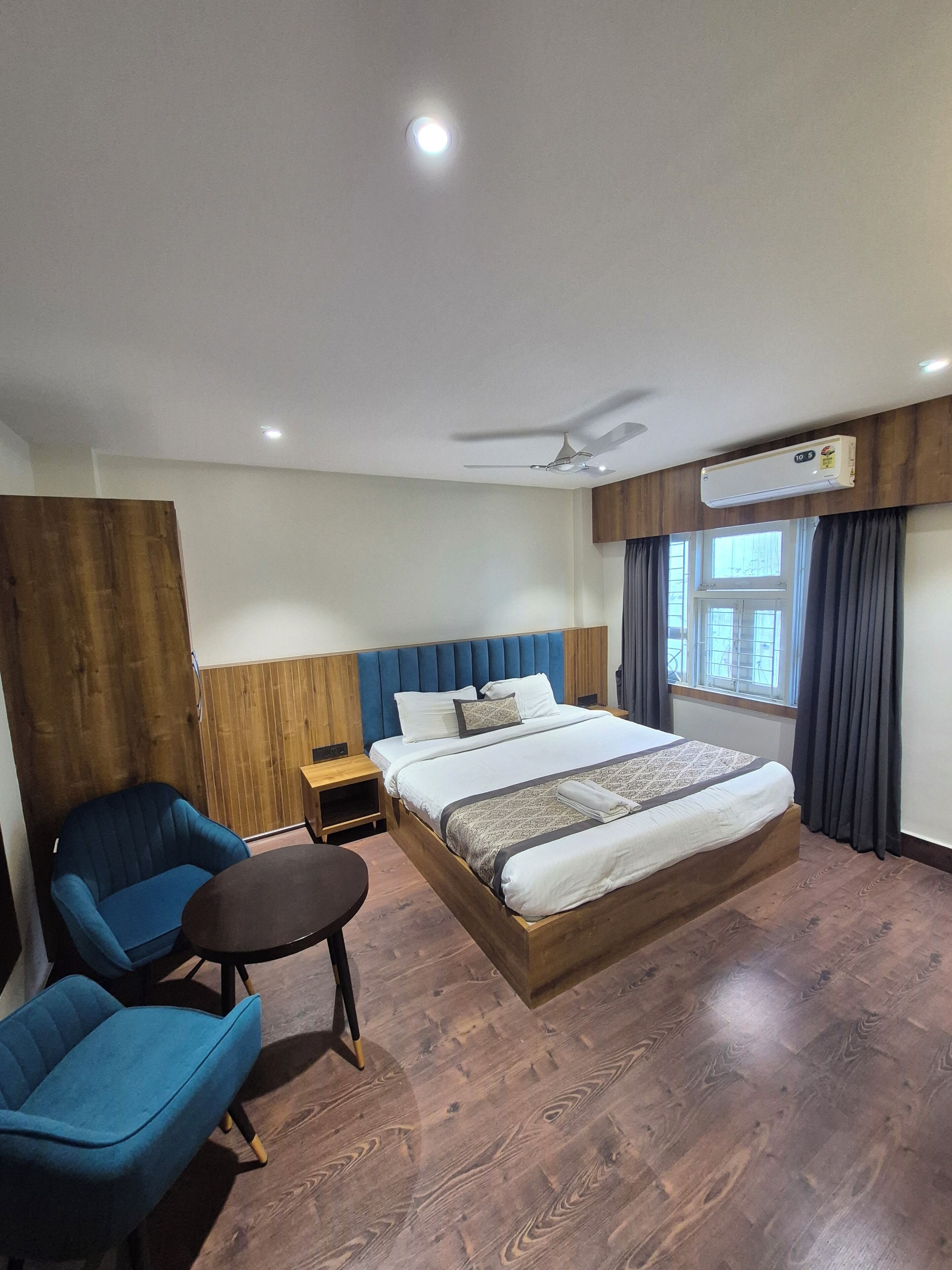 A hotel room with a wooden headboard bed, a blue armchair, a small wooden table, and an air conditioning unit on the wall.