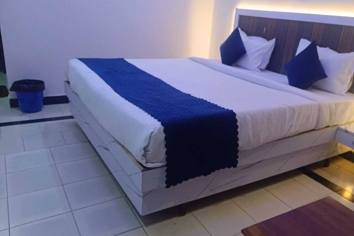 A neatly made bed with a blue throw blanket, white bedding, and two blue pillows. There is a small bedside table with a lamp, a trash bin, and a wall socket.