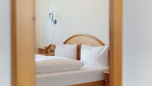 Una cama bien hecha con sábanas blancas y cabecera de madera, una mesita de noche con lámpara, y un aplique en la pared.
