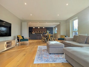 A modern living room with a flat-screen TV, a sofa, a dining area with chairs, and a kitchen in the background.