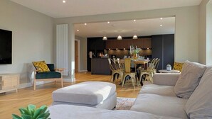A modern living room with a large grey sofa, a dining area with wooden chairs, and a kitchen in the background.