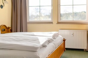 Ein ordentlich bezogenes Bett mit weißen Laken in einem Zimmer mit zwei Fenstern und einer Heizung.