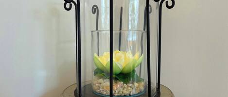 Een transparante glazen vaas met een zwarte metalen standaard, gevuld met een gele bloem en kiezels.