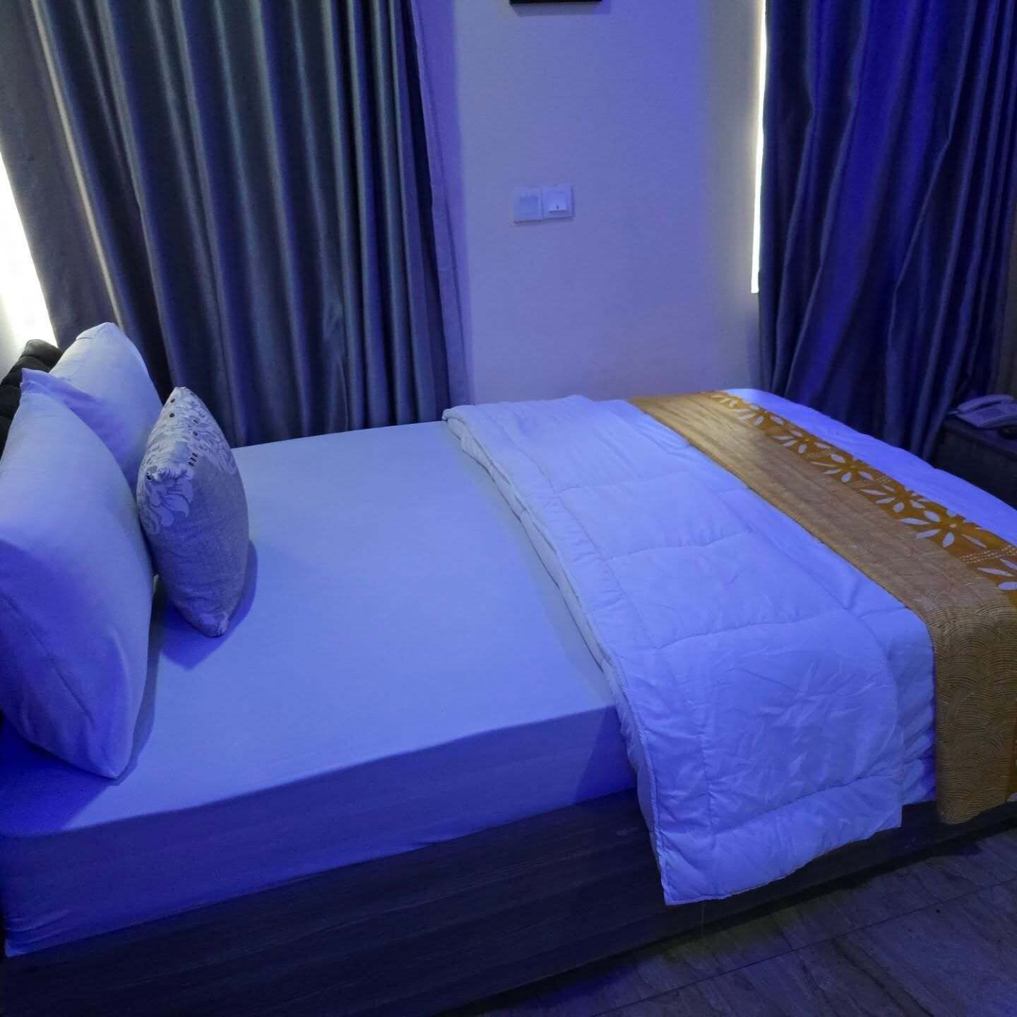 Ein ordentlich bezogenes Bett mit weißen Leinentüchern und einer gemusterten Decke, ein Nachttisch mit einem Telefon und ein Fenster mit Vorhängen.
