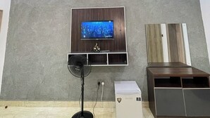 A modern living room with a flat-screen TV mounted on a wall, a standing fan, and a small cabinet. - De Glen Hotel And Suites (Owerri)