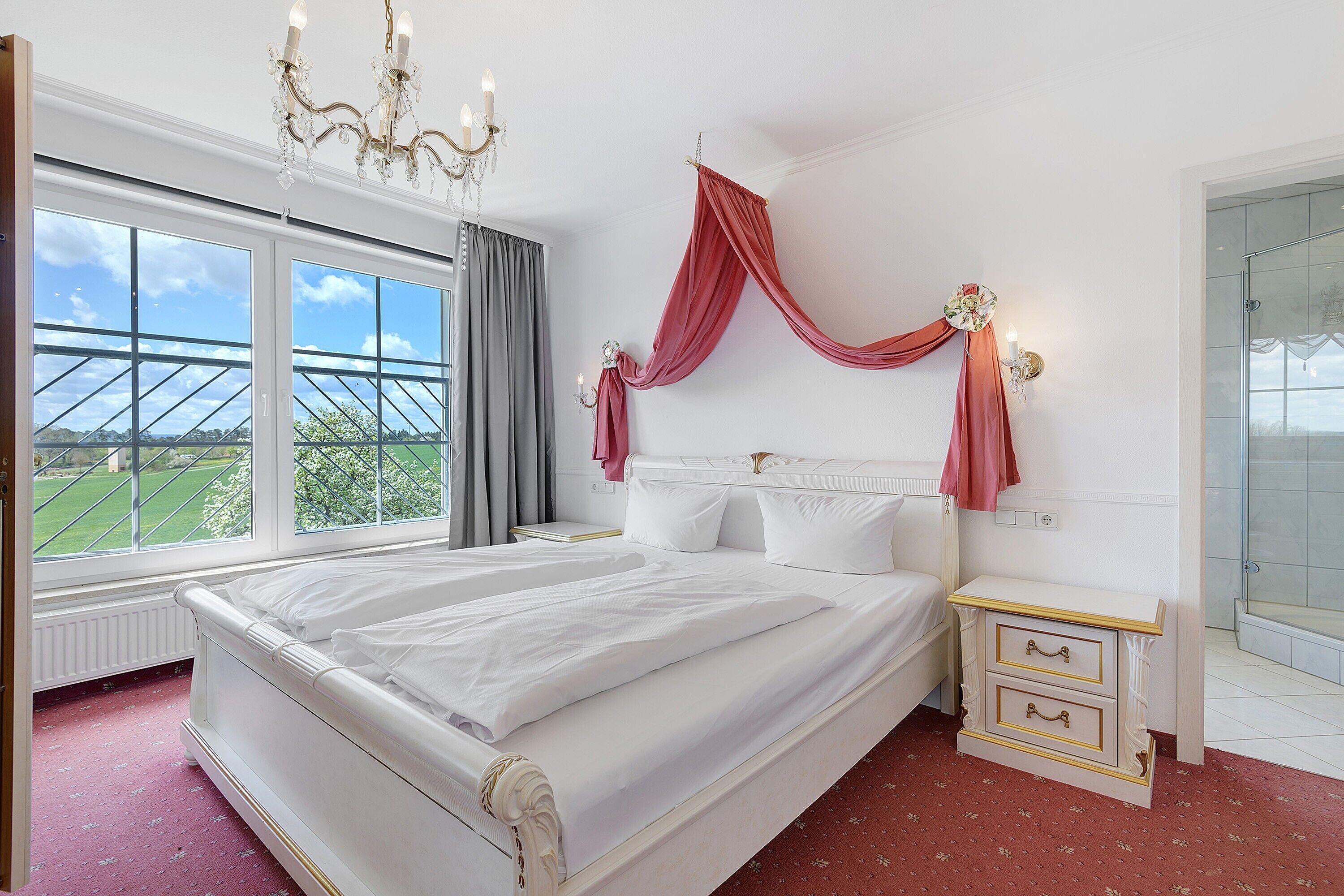 A bedroom with a large bed, a nightstand, a dresser, a chandelier, and a view of the outdoors.