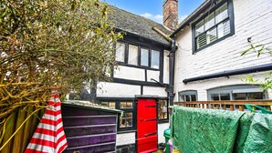 Un callejón estrecho con un edificio blanco, ventanas de marco negro y una puerta roja. Se ve un cobertizo morado a la izquierda, y una lona verde cubre un objeto a la derecha.