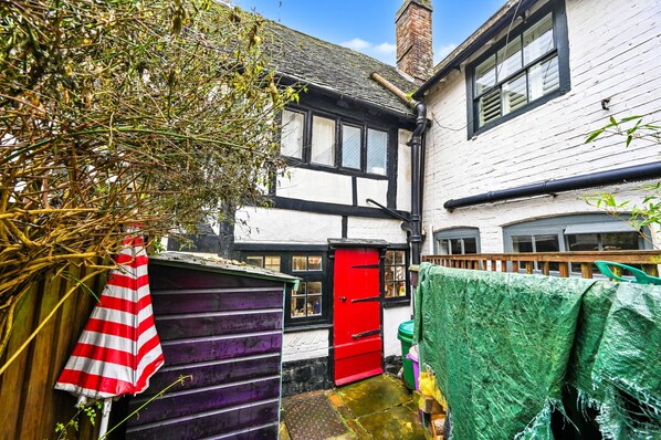 A narrow alleyway with a white building featuring black-framed windows and a red door. A purple shed is visible to the left, and a green tarp covers an object to the right. - Old Town Cottage, Hastings (Hastings)