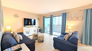 Uma sala de estar com dois sofás, uma mesa de centro, uma TV e uma varanda com vista para edifícios.